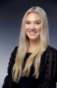 Lady smiling at camera wearing black top, long blonde hair with gray background. Headshot.